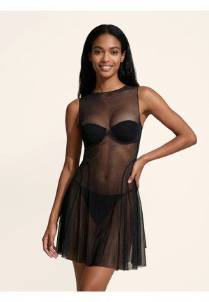 Donna che indossa un vestito nero in rete trasparente senza maniche sopra reggiseno e mutandine nere abbinate, sorridente con la mano sul fianco, su sfondo semplice.