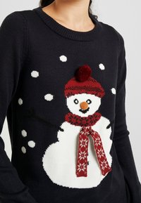 Maglione blu navy con un design di pupazzo di neve bianco, corredato da una sciarpa rossa a motivi, un cappello con pompon e fiocchi di neve bianchi sparsi.