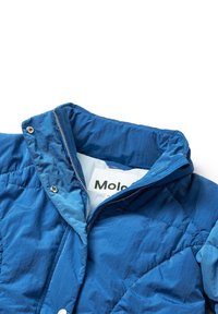 Blå quiltad jacka med hög krage, synlig vit dragkedja och silvrig hårdvara. Har ett texturerat tyg och en märkesetikett inuti.