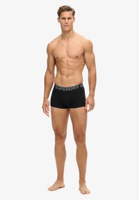 Boxers noirs avec une ceinture logo "SUPERDRY" grise, fabriqués à partir d'un tissu lisse, présentant un design ajusté et sans motifs visibles.
