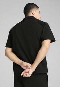 Czarna koszula z krótkim rękawem, o fakturowanej tkaninie, luźnym kroju i płaskim kołnierzyku; widoczne szwy oraz prosta krawędź w talii.