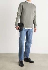 Man i grå sweatshirt, vit skjorta, blå jeans och svarta skor som håller en liten svart väska mot en enfärgad bakgrund.