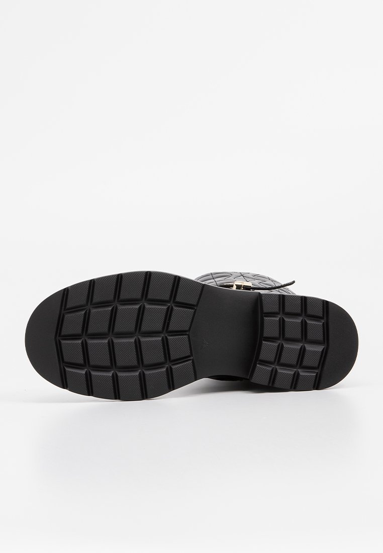 Schwarze Gummisohle mit einer strukturierten, gemusterten Griffigkeit; rechteckige Profilierungen; glatte Oberfläche an den Seiten und ein leichtes Fußgewölbe.