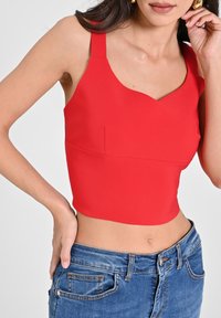 Rotes Crop-Top mit breiten Trägern und einem wellenförmigen Ausschnitt, ausgestattet mit einer glatten Textur und passgenauer Silhouette, kombiniert mit blauen Jeans.