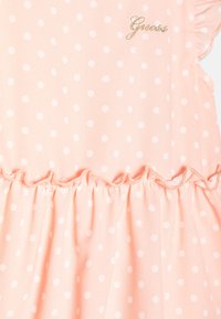 Sukienka w brzoskwiniowe groszki z falbanami na rękawach, wykonana z lekkiego materiału. Posiada złote logo "Guess" na górze oraz zgromadzony w talii.