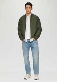Olivengroene bomberjack met drukknoopsluiting, gedragen over een wit gestreept shirt. Blauwe relaxed-fit jeans en beige sneakers maken de outfit compleet.