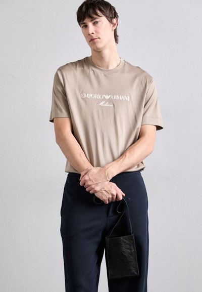 Šviesiai kreminės spalvos medvilnės marškinėliai su apvaliu kaklu ir siuvinėtu "Emporio Armani Milano" logotipu, derinami su tamsiai mėlynomis kelnėmis ir mažu juodu odiniu krepšiu.