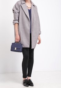 Grå oversized kappa med uppvikta ärmar, lager-på-lager över en marinblå blus, matchad med svarta slim fit-byxor och svarta loafers. Inkluderar en liten blå handväska.