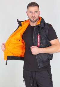 Melns bez piedurknēm veste ar oranžu odere, aprīkota ar rāvējslēdzēja kabatām un birku. Ārējā materiāla virsma ir gluda; iekšējā tekstūra ir kviltota.