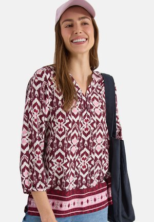 Smilende kvinde iført en lyserød mønstret bluse, lyserød kasket, blå jeans og bærende en mørkeblå skuldertaske.