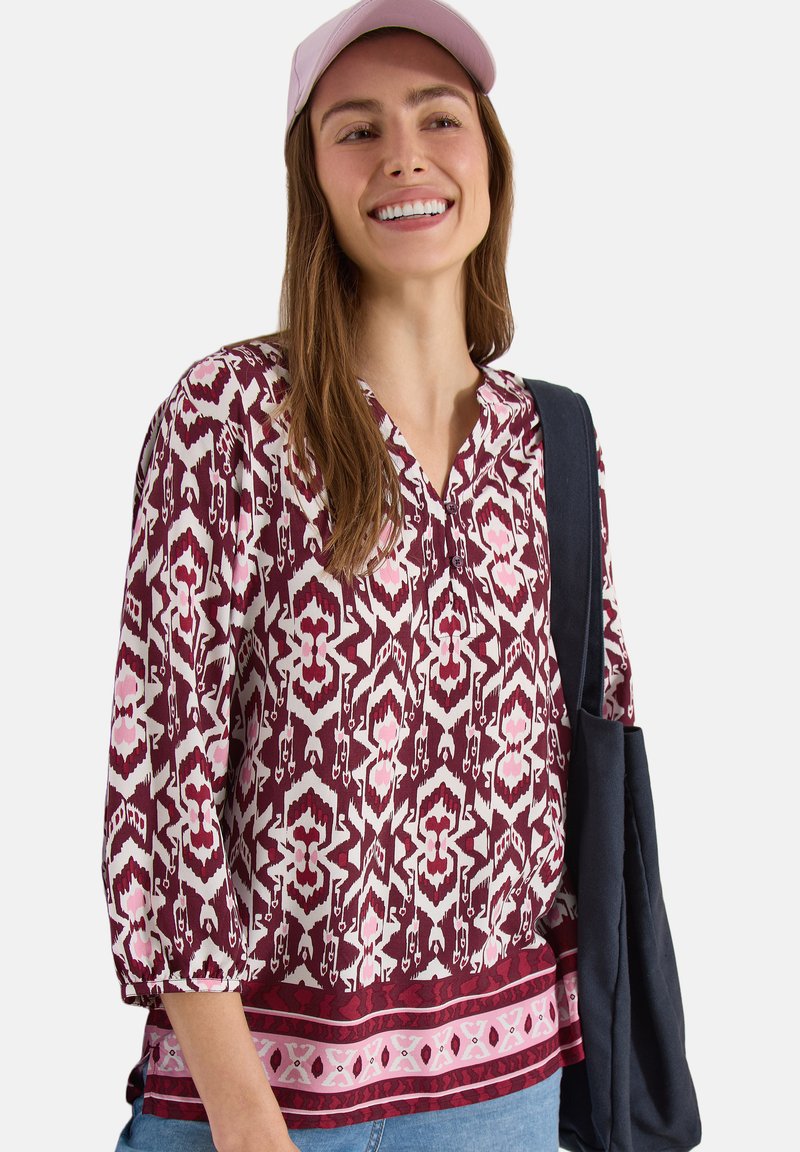 Femme souriante portant un chemisier rose à motifs, une casquette rose clair, un jean bleu et un sac fourre-tout bleu foncé sur l’épaule.