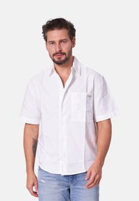 Camisa de algodón blanca de manga corta con un cierre de botones en la parte delantera, un bolsillo en el pecho con una marca y un corte relajado. Textura ligera.