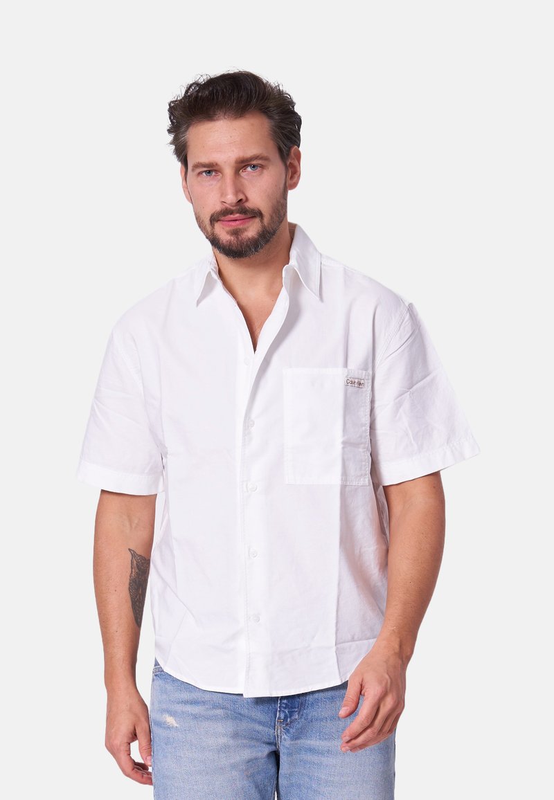 Camisa de algodón blanca de manga corta con un cierre de botones en la parte delantera, un bolsillo en el pecho con una marca y un corte relajado. Textura ligera.