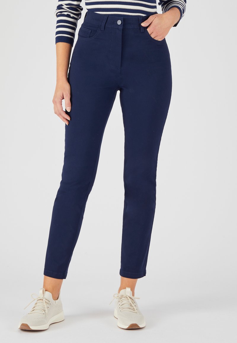 Donkerblauwe high-waisted broek met een slim fit, voorzien van vijf zakken en een knoopsluiting, gecombineerd met lichtgekleurde sneakers met textuur.