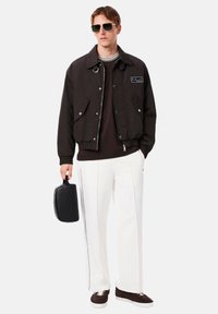 Giacca bomber nera con dettagli in argento, camicia bianca, pantaloni bianchi con strisce laterali, sneakers marroni e una borsa nera. Accessori includono occhiali da sole.