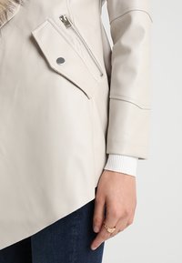 Chaqueta de cuero crema con un bolsillo con cremallera, que presenta una superficie texturizada. Los puños son de canalé blancos, con un detalle de botón a presión plateado visible.