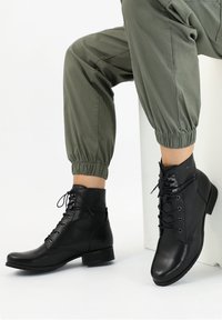Stivali alla caviglia in pelle nera con punta rotonda, lacci anteriori e tacco basso a blocco. Indossati con pantaloni verdi a risvolto. La texture è liscia con dettagli minimi.