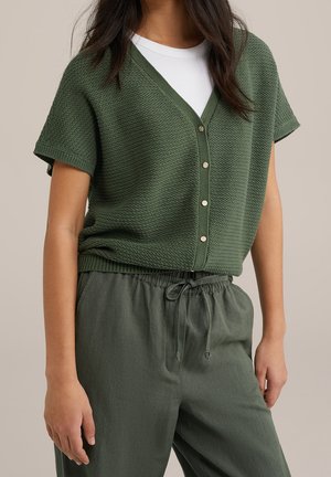 Modèle portant un cardigan texturé à manches courtes et à boutons, vert olive, sur un haut blanc, avec un pantalon assorti à cordon de serrage.