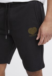 Schwarze Baumwollshorts mit seitlichen Taschen und Kordelzug in der Taille. Mit goldfarbenem Grafik-Design nahe dem Saum, das "CREATE TOMORROW" zeigt.
