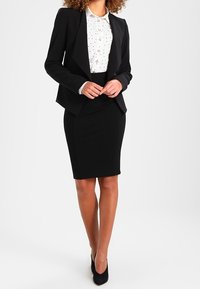 Zwarte, op maat gemaakte blazer over een witte blouse met zwarte polka dots, gecombineerd met een nauwsluitende zwarte kokerrok en zwarte hoge hakken.