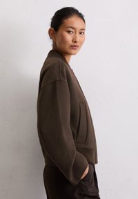 Pull en gros volume marron avec un col et un ourlet côtelés, présentant une poche frontale et une texture lisse, présenté sur un mannequin de profil.