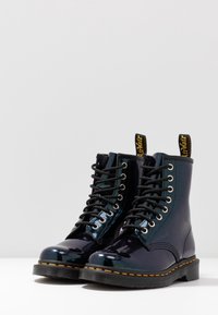 Bottes de combat en cuir verni noir avec une finition brillante, dotées d'œillets en métal argenté, de lacets noirs et d'une couture de semelle orange contrastante.