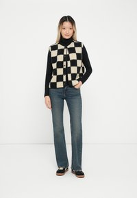Gilet a scacchi nero e crema con bottoni, indossato su un dolcevita nero, abbinato a jeans blu e scarpe nere. Sfondo bianco semplice.