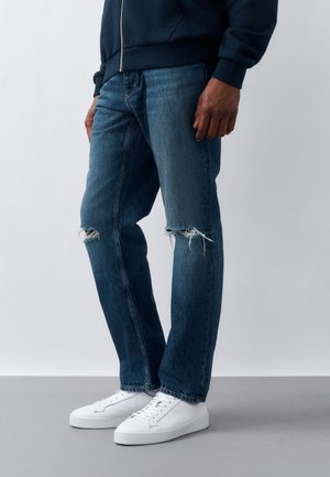 Muž oblečený v tmavomodrých roztrhaných džínsoch, bielych teniskách a tmavomodrej bunde na zips stojaci pred jednofarebným pozadím.