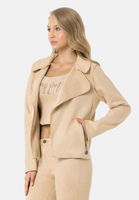 Veste de moto en suédine beige avec un large col, des poches zippées et une silhouette ajustée. Accompagnée d'un haut court assorti.