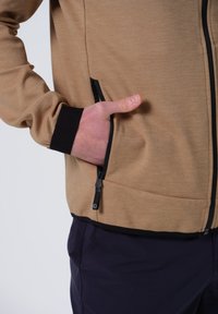 Chaqueta de cremallera de color marrón claro con detalles en negro, que cuenta con un bolsillo lateral y una tela texturizada. Las mangas son negras, contrastando con el color del cuerpo.