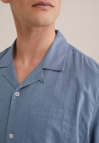 Camisa de manga corta azul claro, hecha de una tela suave, con un cuello notch, dos botones y un bolsillo en el pecho con un acabado limpio.