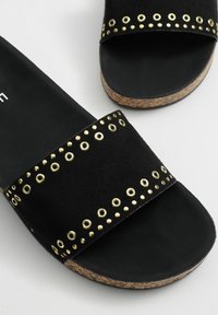 Chanclas negras con una parte superior suave similar al ante, adornadas con detalles de hardware dorados y una suela de corcho. El diseño incluye recortes circulares en la correa.