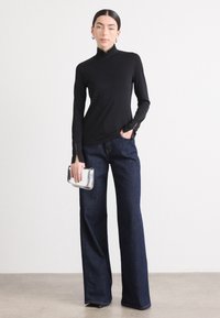 Zwarte lange mouwen top met hoge kraag en ritssluitingen, gecombineerd met brede donkere denim jeans. Model houdt een zilveren clutch vast.
