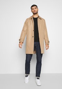 Abrigo trinchera beige con cuello y botones negros, llevado encima de una camiseta negra y combinado con jeans oscuros y zapatillas blancas.
