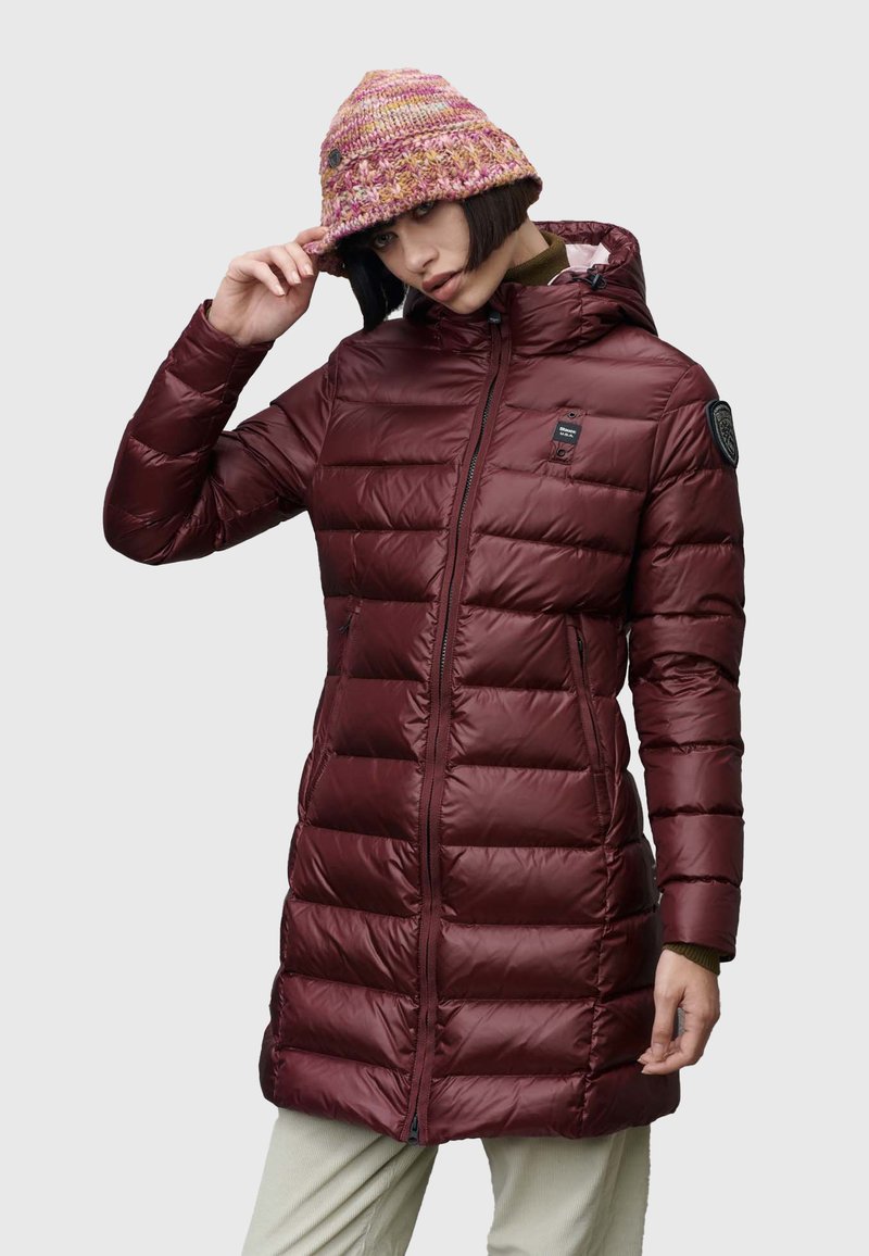 Maroon puffer coat with horizontal baffles, front zipper, and hood. Worn with a multicolored knitted hat, displaying a textured pattern.