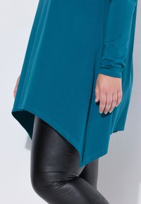 Długi rękaw w kolorze zielonkawoniebieskim z asymetrycznym dołem, zestawiony z błyszczącymi czarnymi legginsami. Tkanina wydaje się miękka o gładkiej fakturze.