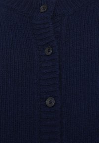 Tuvums skats uz tumši zilu adītu kardigānu ar ribotu struktūru un četriem melniem apaļiem pogām centrā.