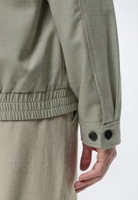 Gros plan du bras d'une personne portant une chemise verte à manches longues avec poignets boutonnés et taille élastique, reposant sur le côté.