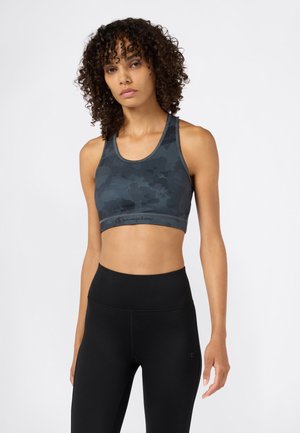 Mujer con cabello rizado lleva un sujetador deportivo de camuflaje azul oscuro y leggings negros de talle alto sobre un fondo claro.
