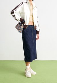 Vit zip-up jacka med bruna accenter, beige crop top, mörk denim midi kjol och vita sneakers med beige accenter. Liten mönstrad handväska.