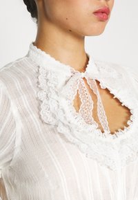Femme portant un chemisier en dentelle blanche avec un col à volants noué en nœud et un décolleté en forme de clé montrant partiellement le cou et les lèvres.