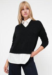 Jersey negro de pico superpuesto sobre una camisa blanca con cuello, que presenta dobladillos acanalados y un ajuste relajado. Tejido fino y suave con una textura sutil.