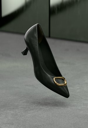 Black leather pointed-toe pump with a small geometric heel and a gold oval buckle accent on the vamp. Smooth texture and sleek design.