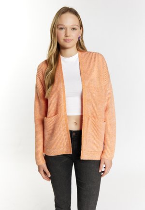 Oranžový pletený cardigan s volným střihem, otevřeným předním dílem a dvěma předními kapsami. Hladký povrch a dlouhé rukávy, nošený přes bílý top.