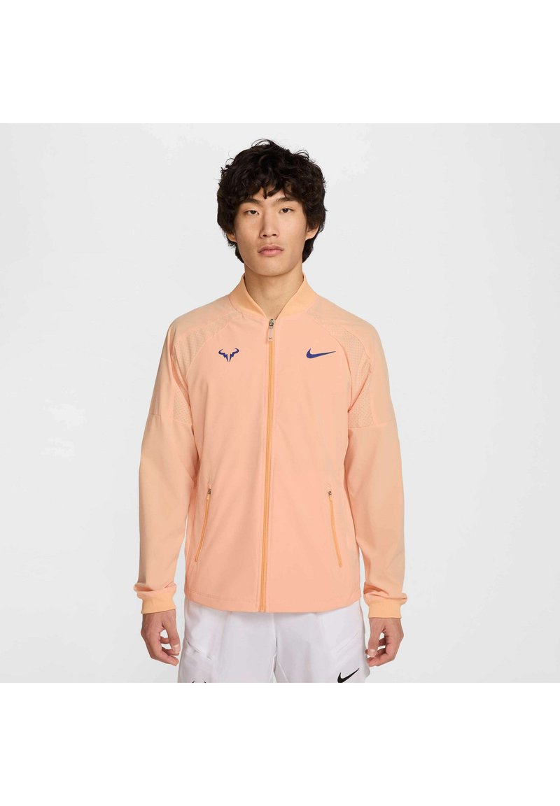 Chamarra zip ligera en color durazno claro con acentos del logotipo de Nike y un toro. Cuenta con dos bolsillos delanteros con zipper y paneles texturizados en las mangas.