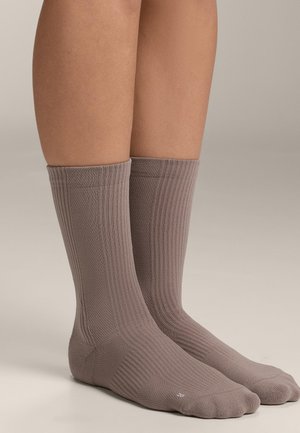 Benen met halflange sokken in taupekleur met een geribbelde textuur op een lichte, neutrale achtergrond, waarbij de pasvorm en stofdetails van de sokken worden getoond.