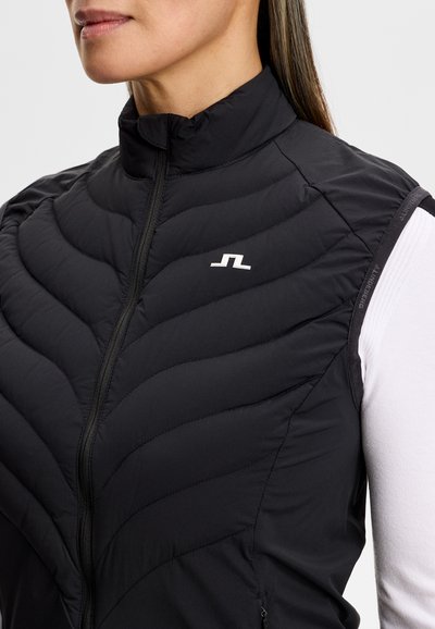 Kvinde iført en sort quiltet ærmeløs vest med et hvidt logo på brystet, over en hvid langærmet skjorte.