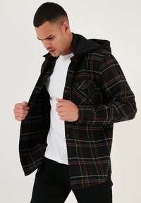 Camicia flanella a quadri nera e rossa con cappuccio nero. Presenta una chiusura frontale con bottoni, tasca sul petto e linee bianche e blu a contrasto nel motivo.