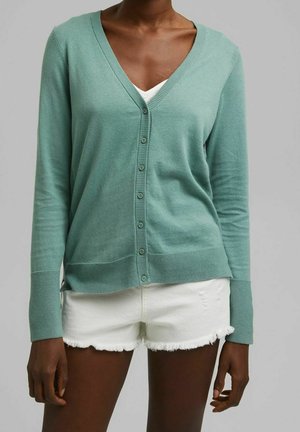 Frau trägt eine helltürkisfarbene, geknöpfte Strickjacke über einem weißen Oberteil, kombiniert mit weißen Shorts mit ausgefranstem Saum vor einem schlichten grauen Hintergrund.