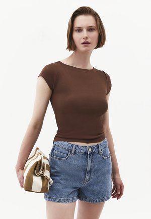 Vrouw met kort bruin haar die een bruin nauwsluitend T-shirt met korte mouwen draagt, blauwe denimshort en een beige-witte gestreepte tas vasthoudt.
